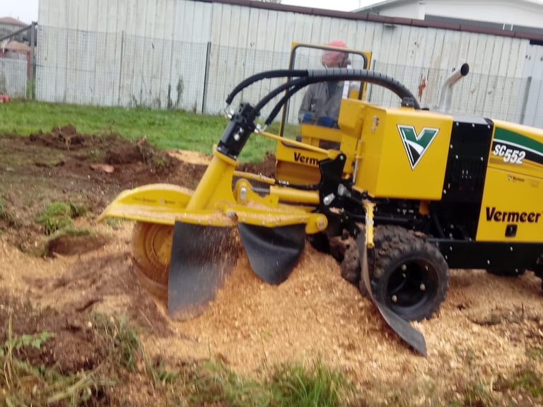 Yellow Vermeer stump grinder cutting into a tree stump on a dirt lot with metal buildings in the background
