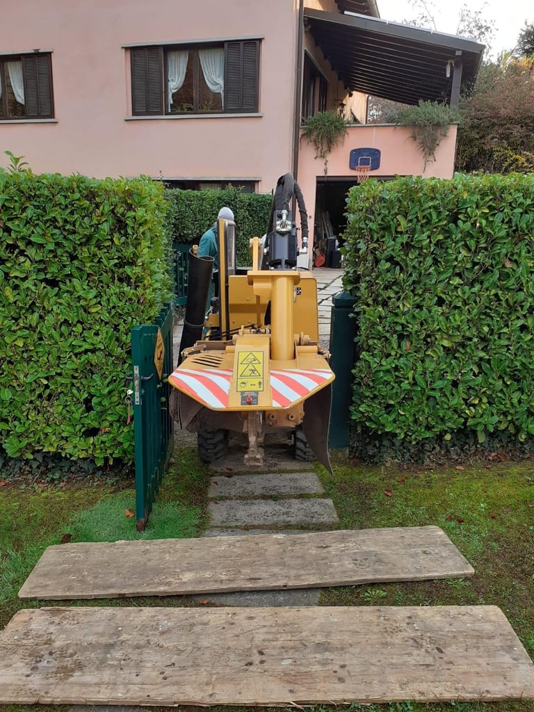 Yellow compact loader parked in a residential driveway between green hedges, leading to a pink house